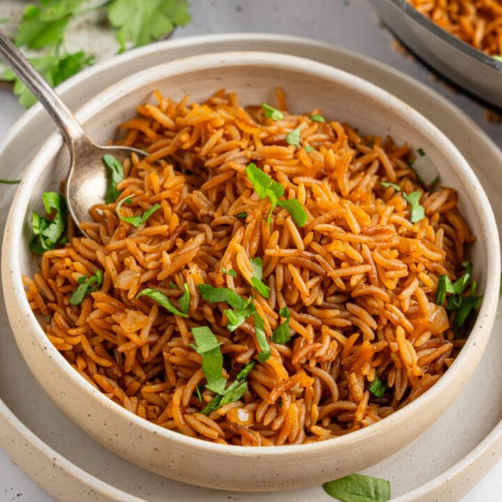 A bowl of homemade Texas Roadhouse seasoned rice garnished with fresh parsley. The bowl is on a ceramic plate with a spoon. The plate is on a white counter with a skillet of rice and small ramekin of garnishes.