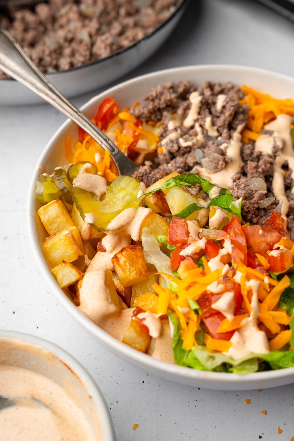 A loaded burger bowl with shredded lettuce, ground beef, roasted potatoes, and sliced vegetables with a loaded burger sauce. There is a fork in the bowl. The bowl is on a white counter with a skillet of ground beef and ramekin of burger sauce.