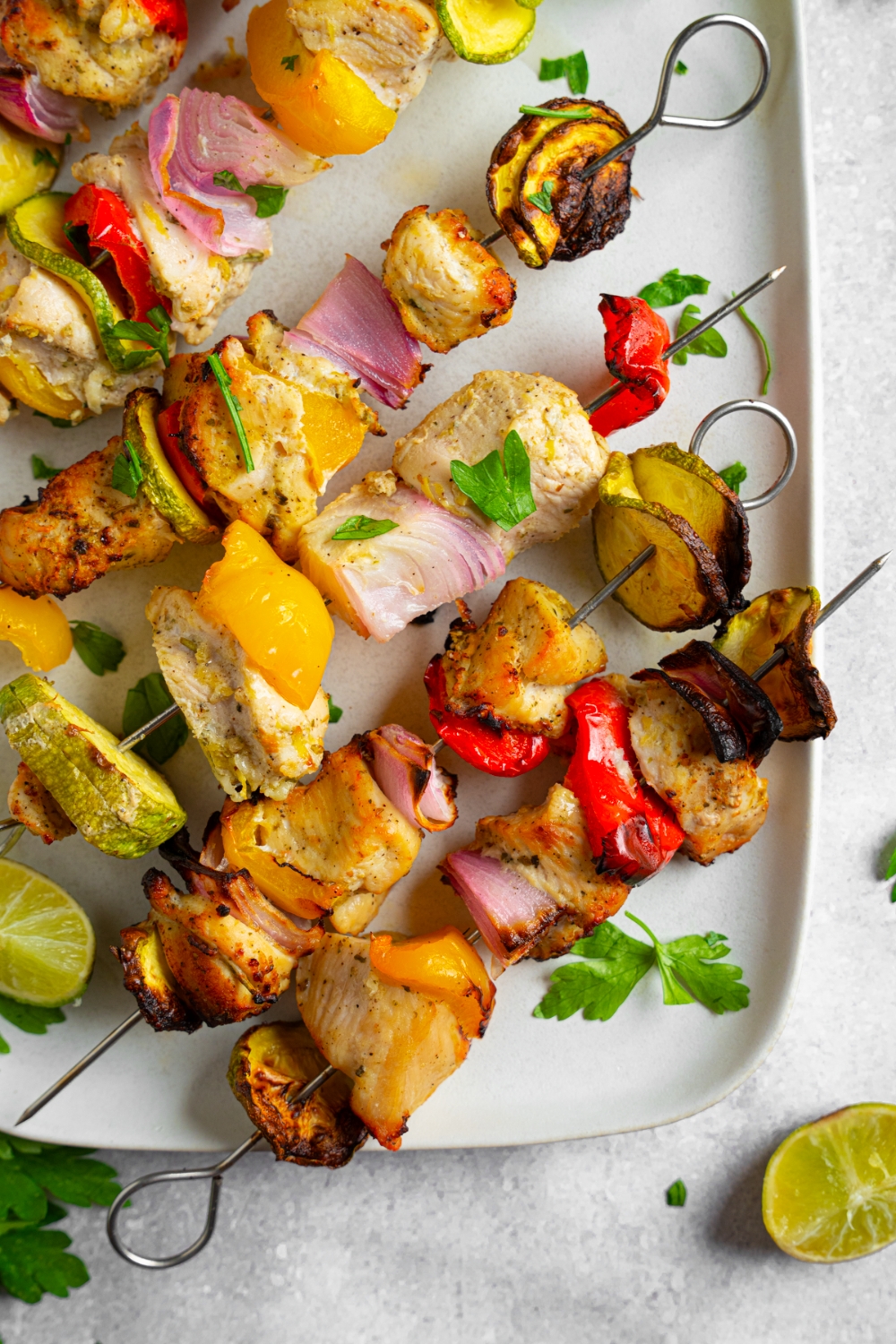 Six oven roasted chicken kebabs consisting of chicken, red onion, red bell pepper, yellow bell pepper, and zucchini garnished with lime. The kebabs are on a white plate garnished with fresh cilantro.