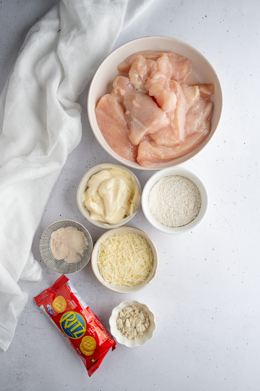 An overhead shot of several bowls in various sizes containing ingredients to make crack chicken including flour, chicken, mayo, parmesan, ranch seasoning, ritz crackers, and seasonings.