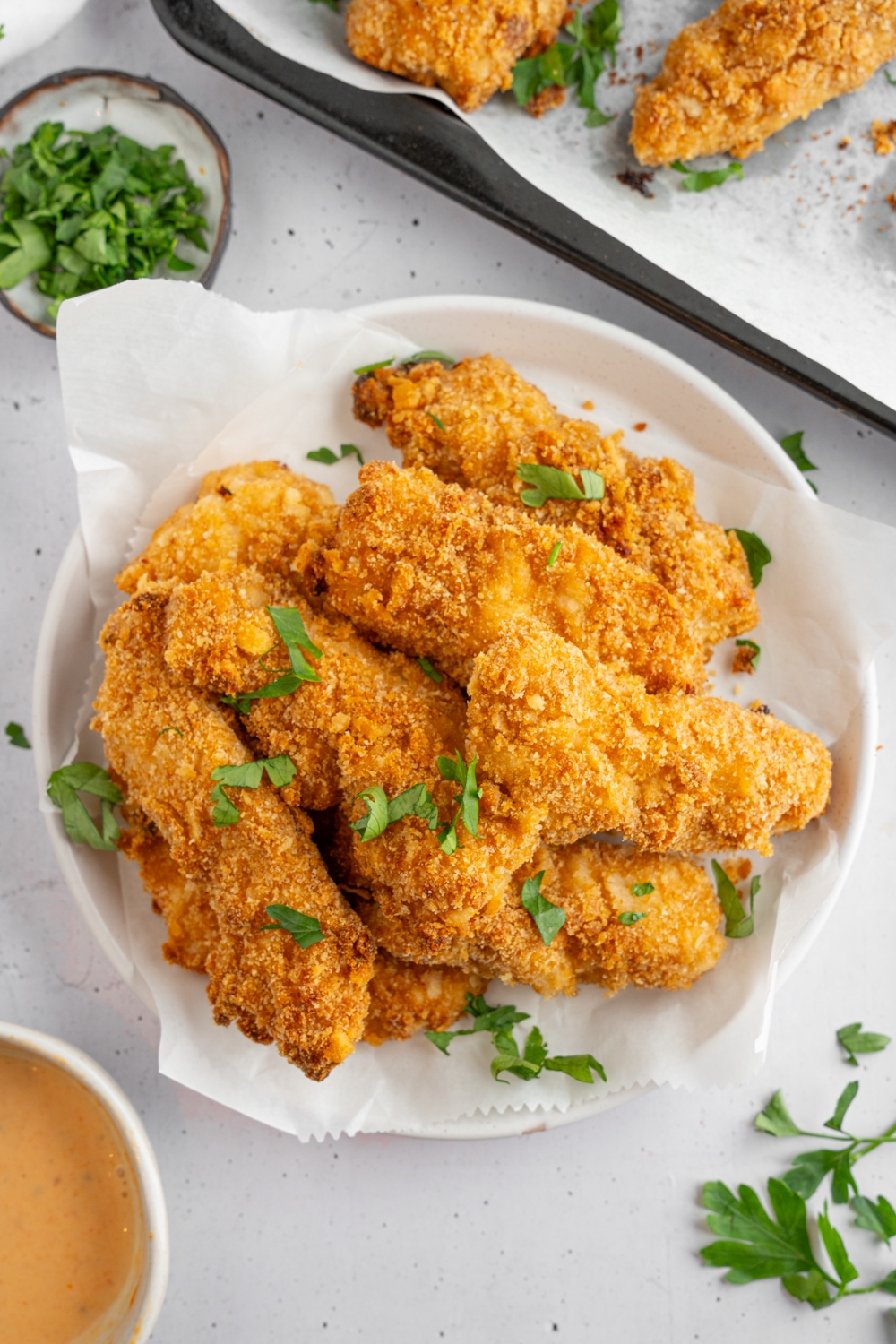 Crack chicken garnished with fresh parsley served in a bowl lined with parchment paper. The bowl is on a white counter with a tray of chicken and small ramekin of garnishes and bowl of dipping sauce.