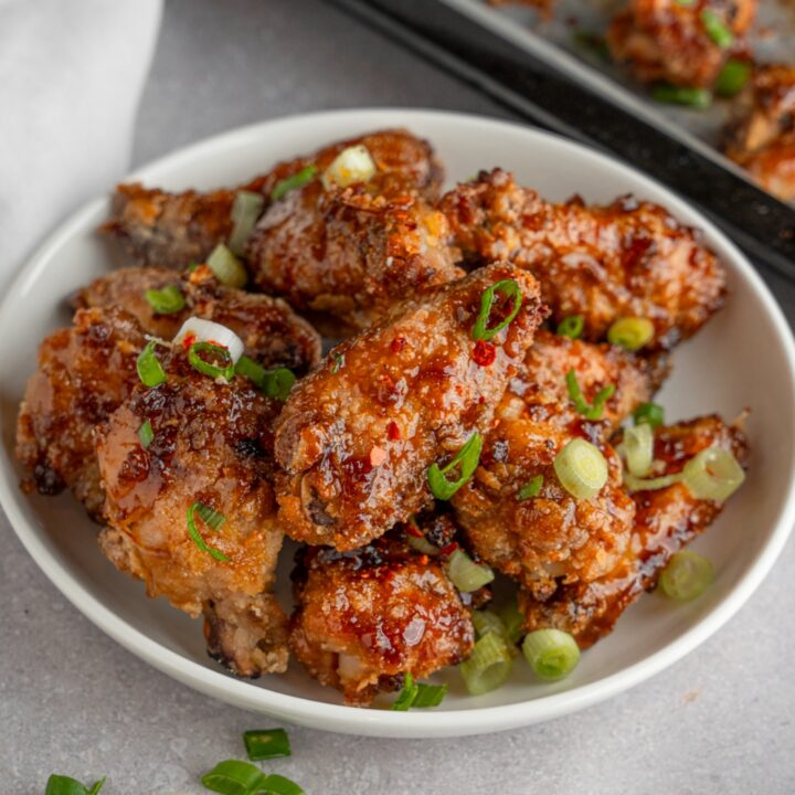 A bowl of fish sauce chicken wings garnished with sliced green onion. The bowl is on a white counter with a baking sheet of wings and small ramekin of garnishes.