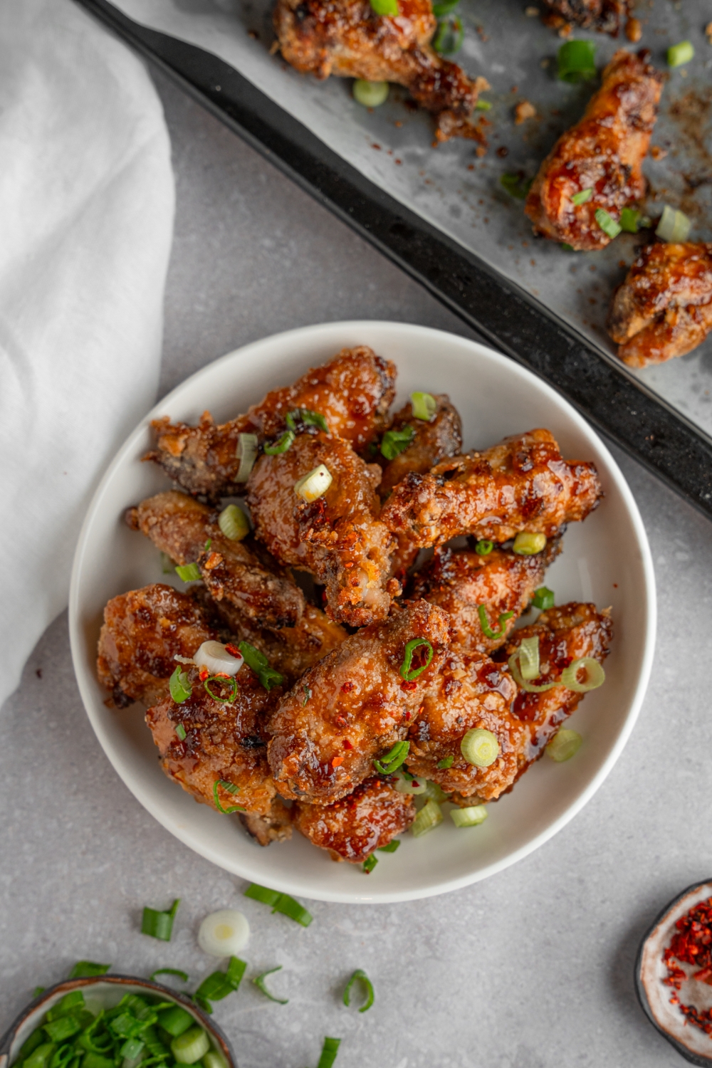A bowl of fish sauce chicken wings garnished with sliced green onion. The bowl is on a white counter with a baking sheet of wings and small ramekin of garnishes.