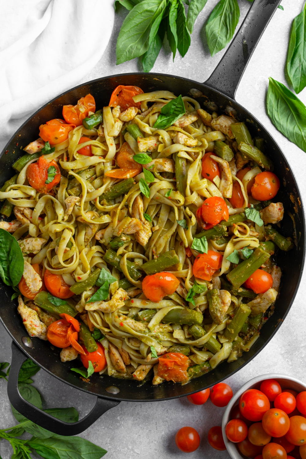 A skillet with fettuccine pasta tossed with asparagus, chicken, and tomatoes in a pesto sauce garnished with fresh basil. The skillet is on a white counter with a small bowl of cherry tomatoes.