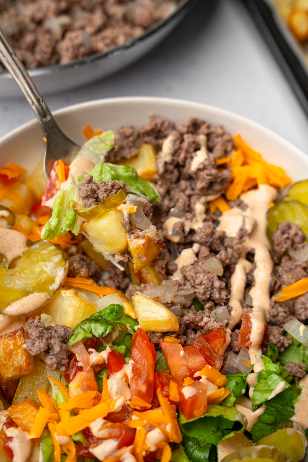 A fork taking a bite from a loaded burger bowl. The bowl consists of ground beef, roasted potatoes, shredded lettuce, sliced vegetables, and loaded burger sauce. The bowl is on a white counter with. a pan of ground beef.
