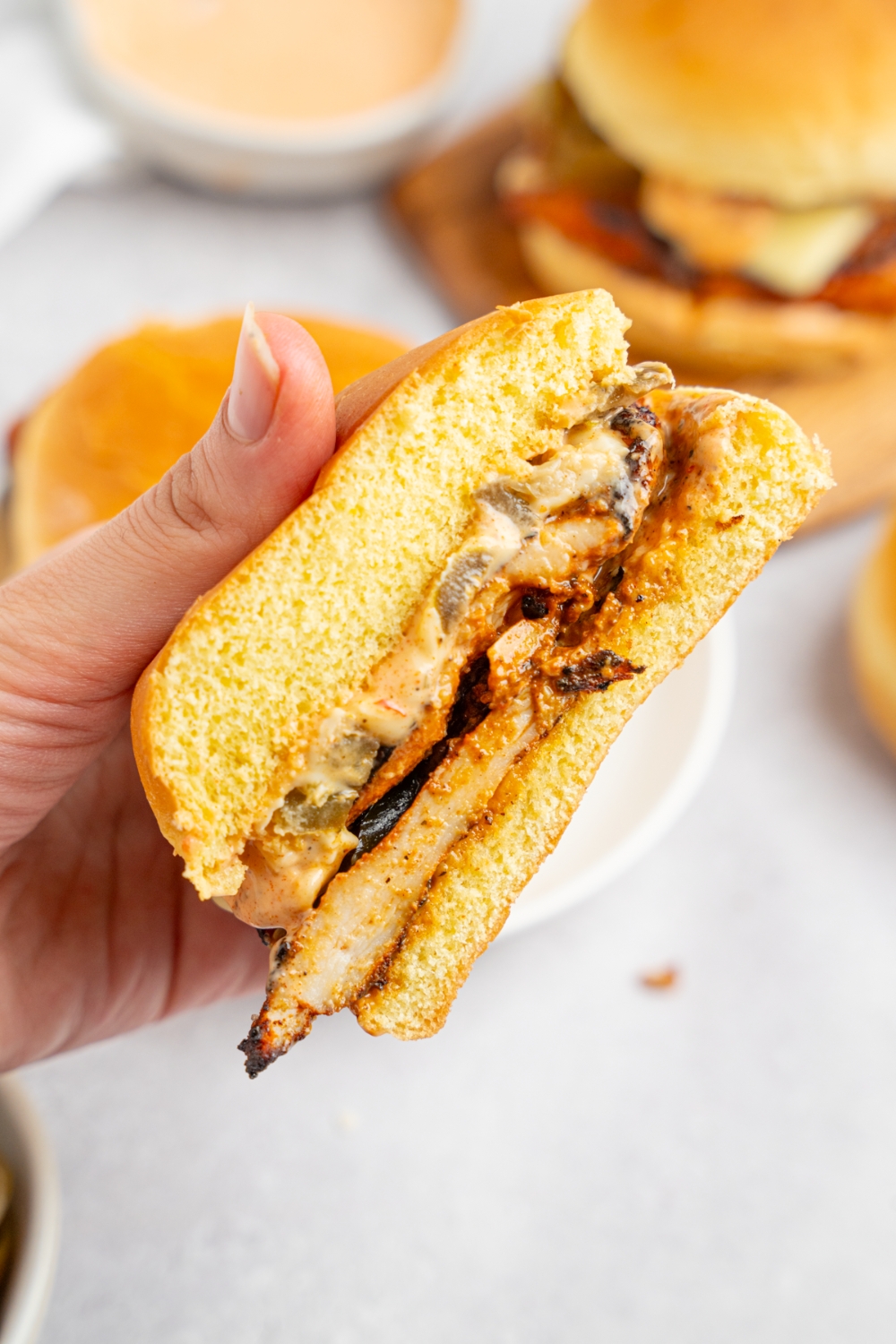 A hand holding a homemade Popeye's blackened chicken sandwich sliced in half. There is a plate of sandwiches on a white counter blurred in the background.
