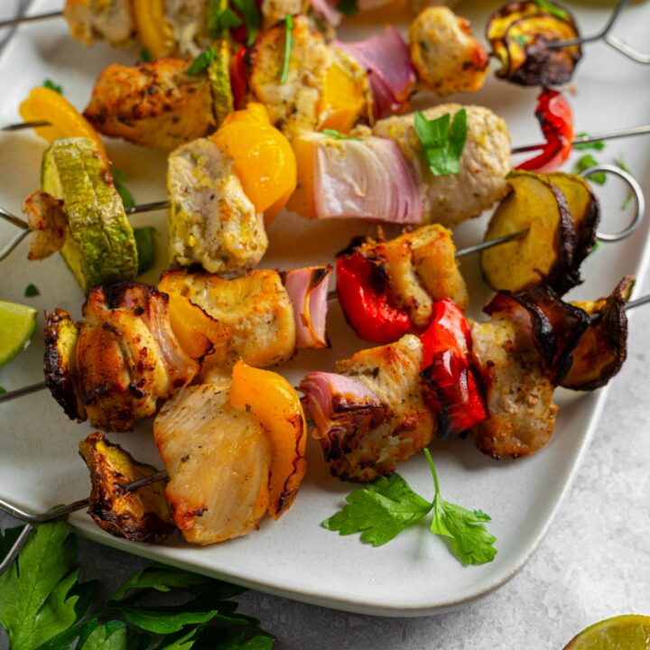 Six oven roasted chicken kebabs consisting of chicken, red onion, red bell pepper, yellow bell pepper, and zucchini garnished with lime. The kebabs are on a white plate garnished with fresh cilantro.