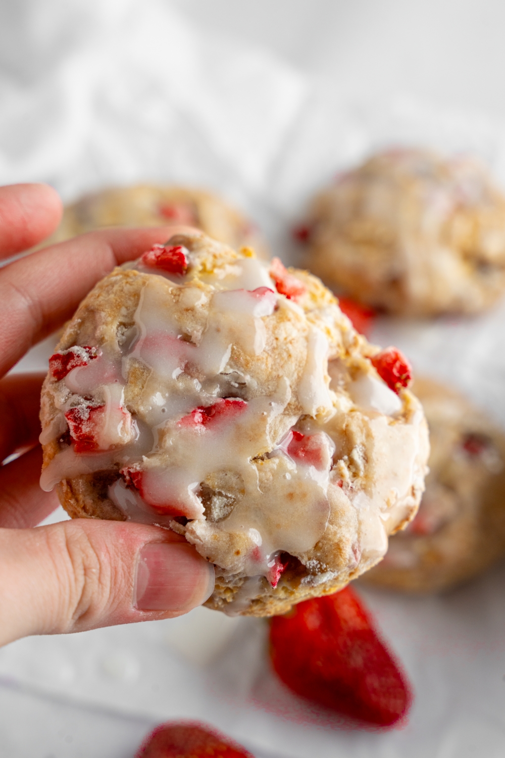 A close up of a hand holding a homemade Popeye's strawberry biscuit. There are additional biscuits on a piece of parchment paper blurred in the background.