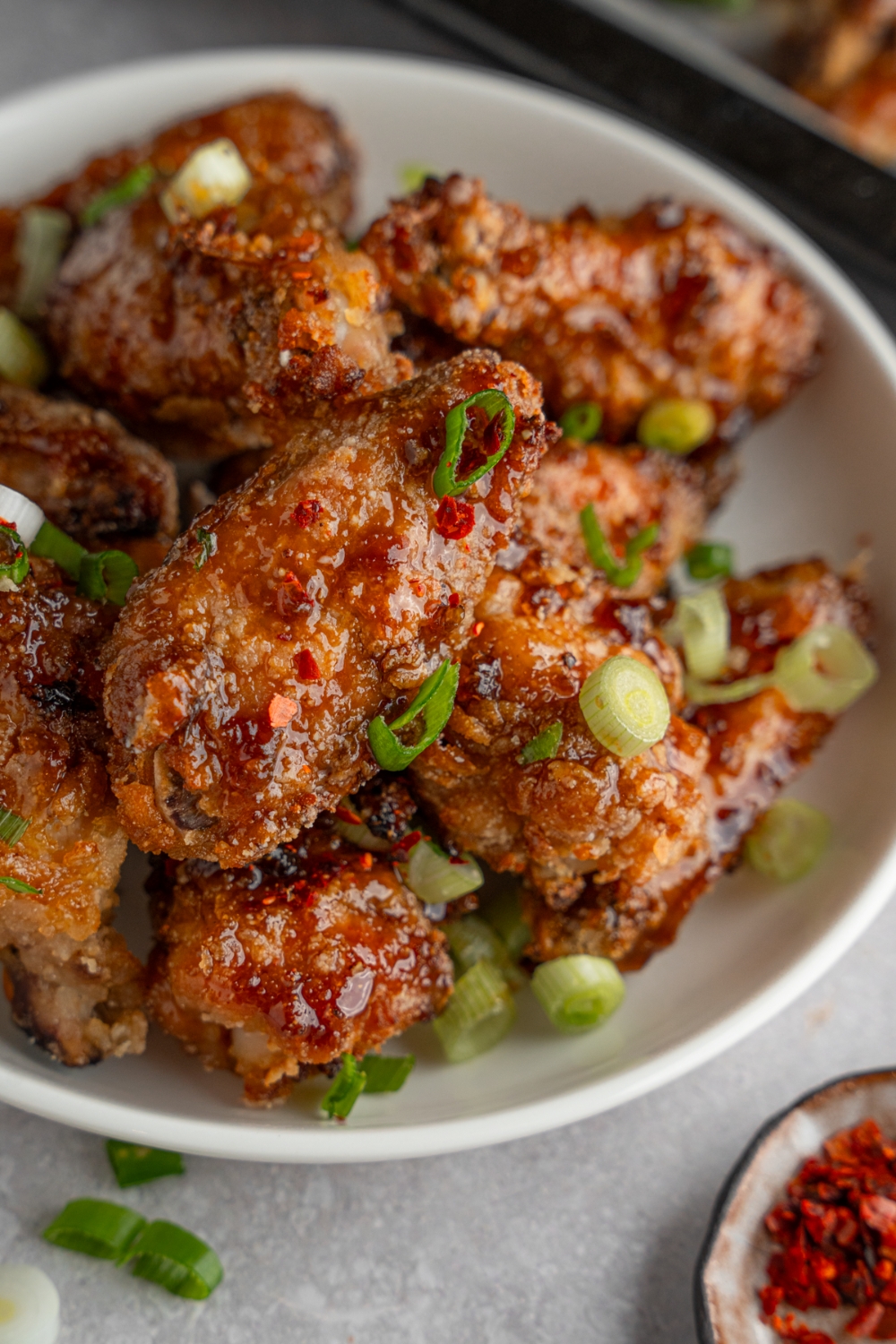 A bowl of fish sauce chicken wings garnished with sliced green onion. The bowl is on a white counter with a baking sheet of wings and small ramekin of garnishes.
