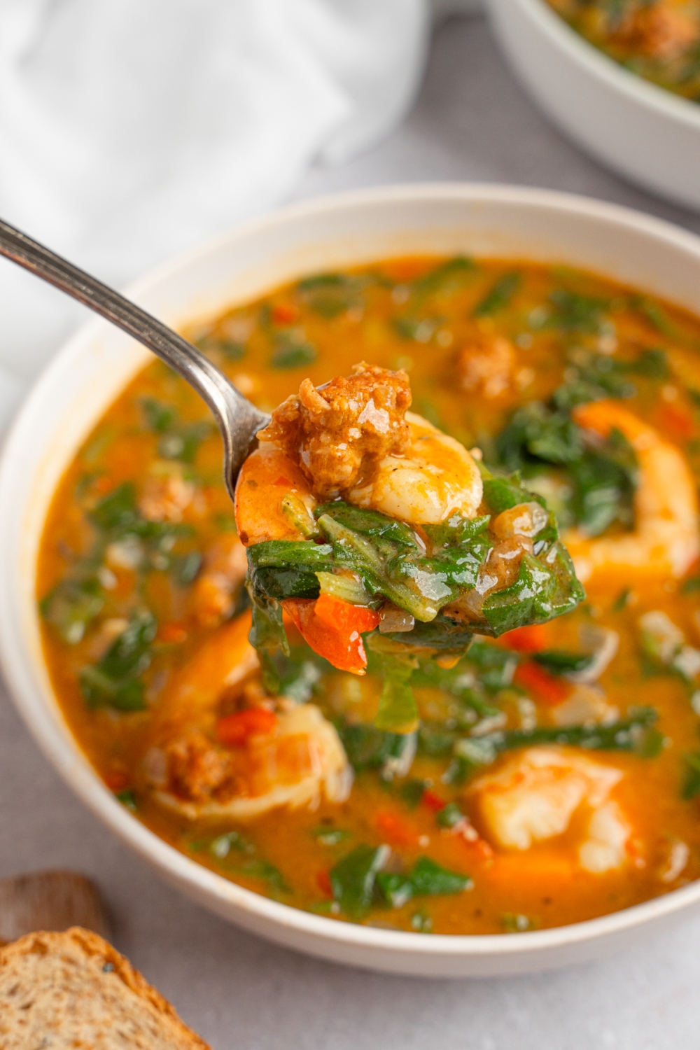 A close up of a spoon with a bite of gumbo greens. There is a bowl of gumbo on a white counter with an additional bowl of gumbo blurred in the background.