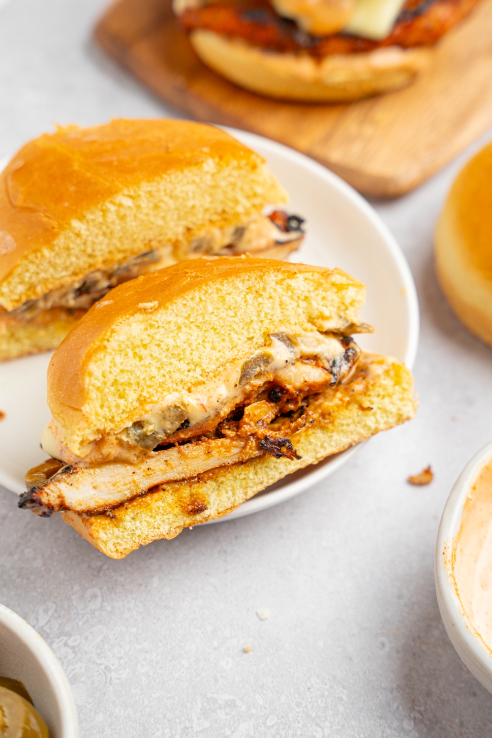 A homemade Popeye's blackened chicken sandwich sliced in half on a white plate. The plate is on white counter with an additional sandwich on a wooden board.