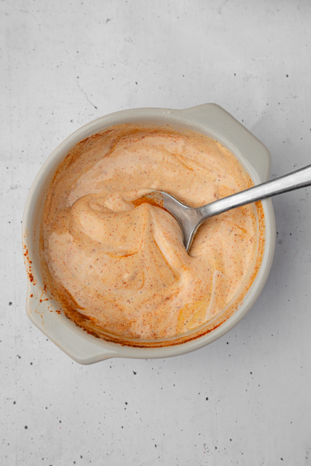 A small ramekin with a spoon mixing a loaded burger sauce. The ramekin is on a white counter.