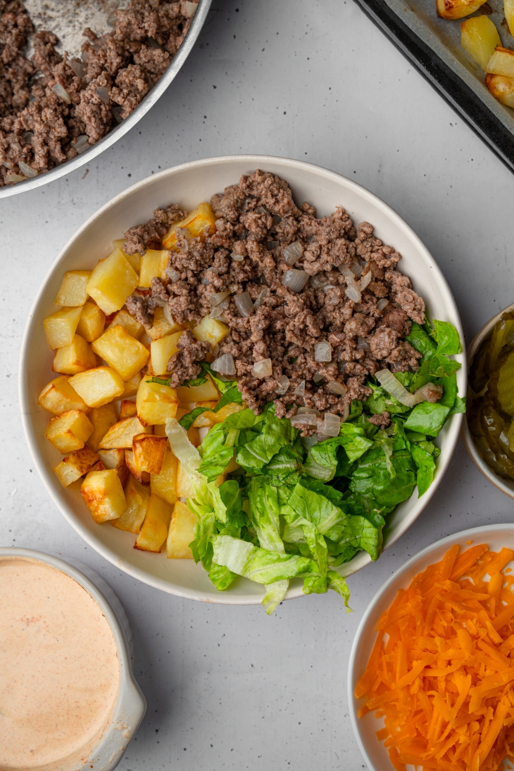 A white salad bowl with shredded lettuce, ground beef and onions, and roasted potatoes. The bowl is on a white counter with a bowl of shredded carrot, pan with ground beef, and ramekin of loaded burger sauce.