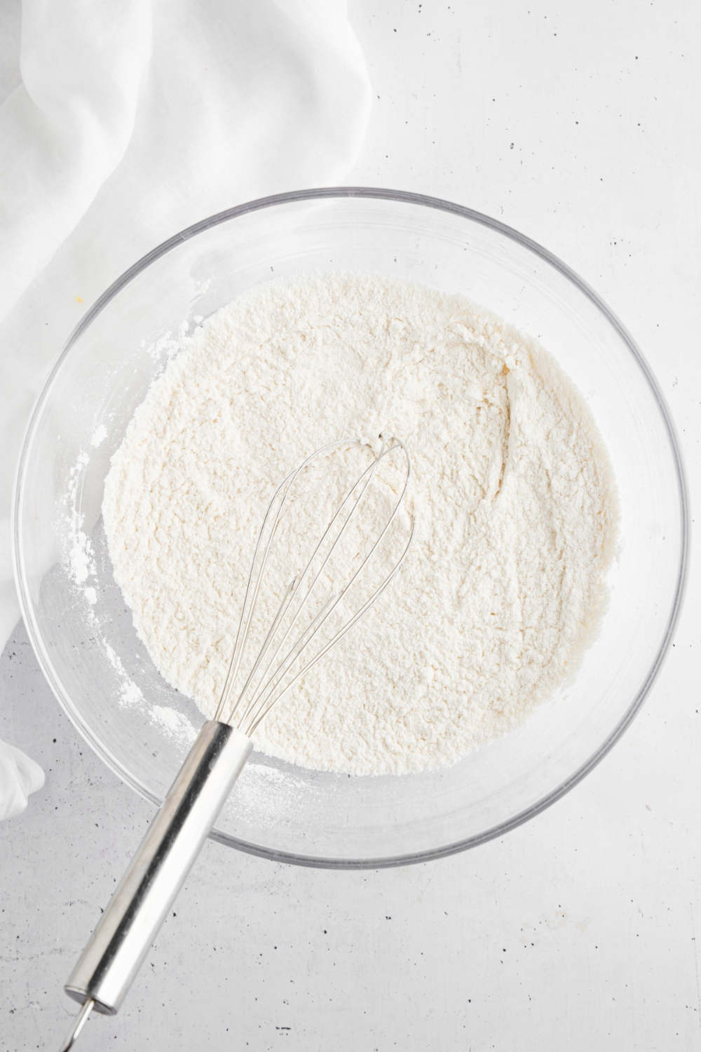 A glass bowl with a whisk combining dry ingredients for dough. The bowl is on a white counter with a white cloth napkin.
