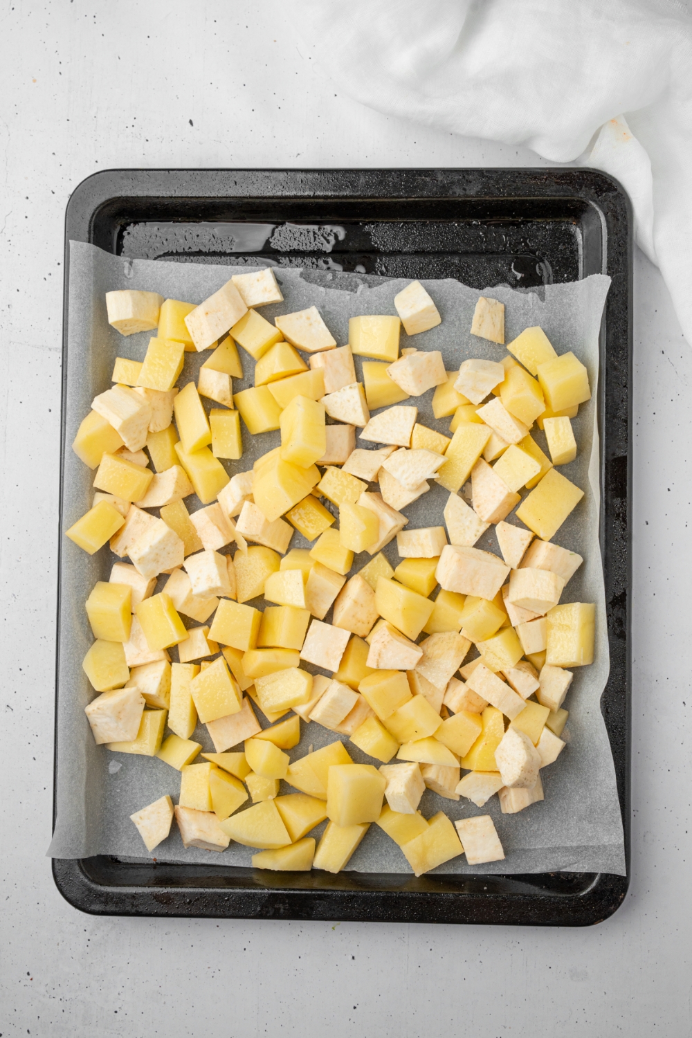 A baking tray lined with parchment paper with diced potatoes. The tray is on a white counter.