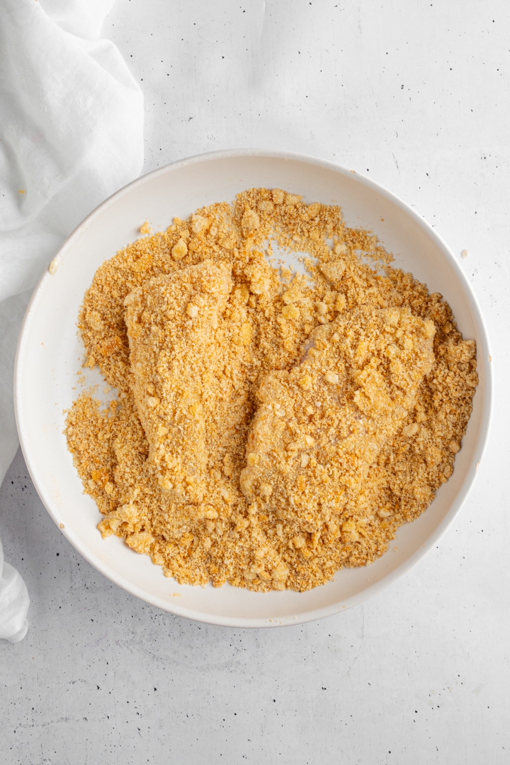 A bowl with two pieces of chicken coated in crumbled ritz crackers. The bowl is on a white counter.