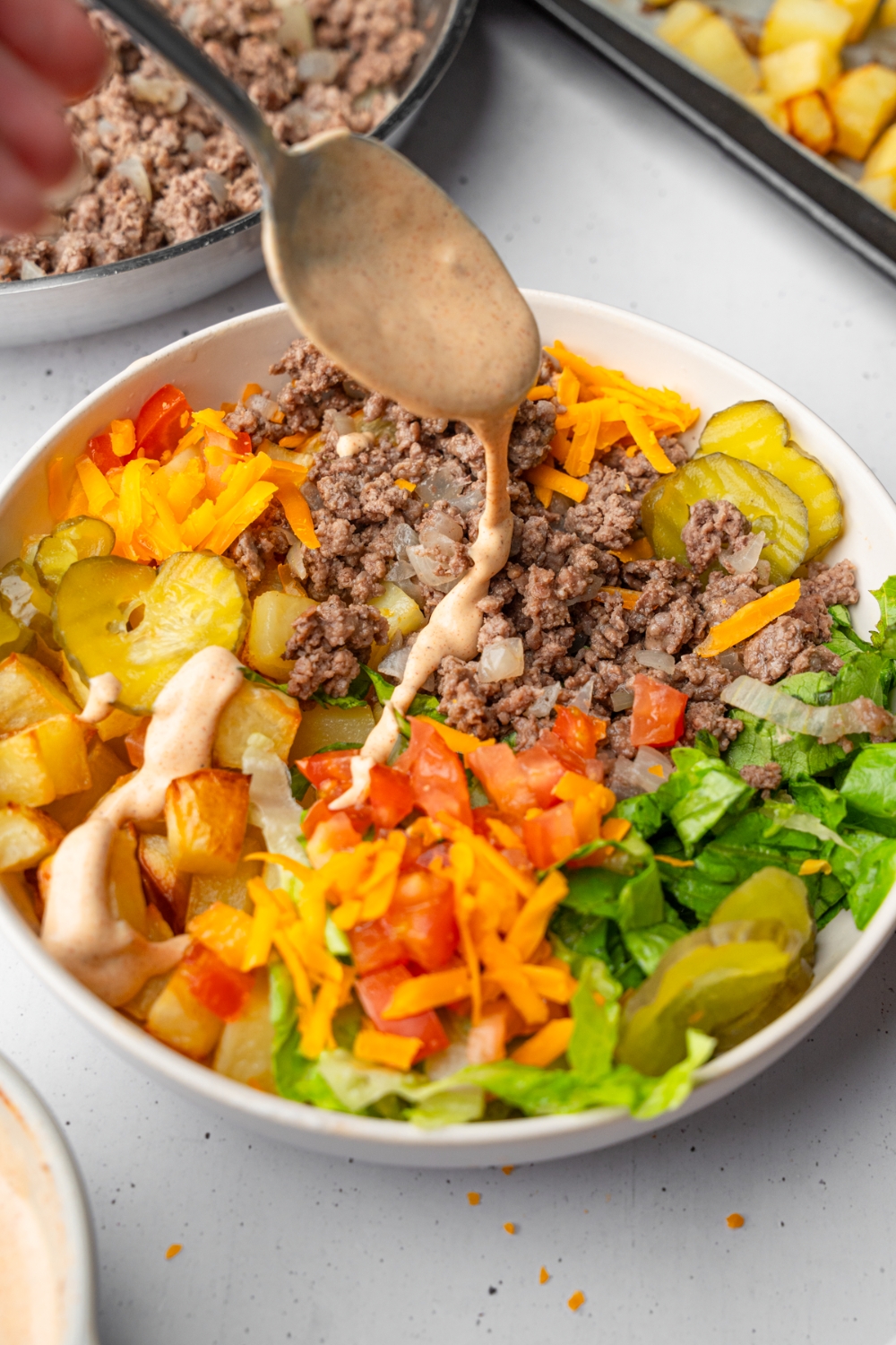 A loaded burger bowl with ground beef, roasted potatoes, shredded lettuce, and sliced vegetables. There is a spoon drizzling a loaded burger sauce over the salad. The bowl is on a white counter with a skillet of ground beef and tray of roasted potatoes.