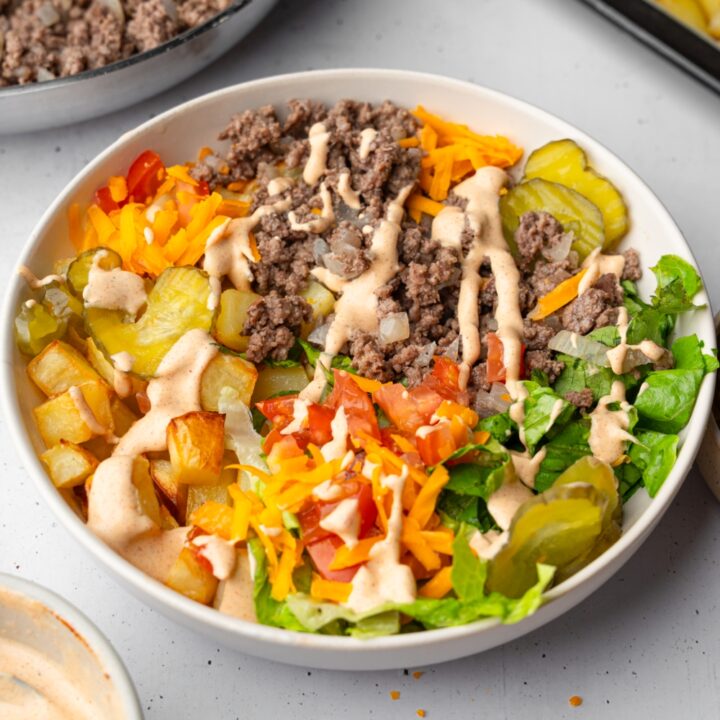 A loaded burger bowl with shredded lettuce, ground beef, roasted potatoes, and sliced vegetables with a loaded burger sauce. The bowl is on a white counter with a skillet of ground beef and tray of roasted potatoes.