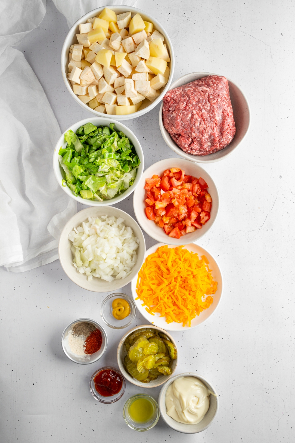 An overhead shot of several bowls in various sizes containing ingredients to make loaded burger bowls including shredded lettuce, diced potatoes, ground beef, diced onions, tomatoes, shredded cheese, pickles, mayo, ketchup, and more.