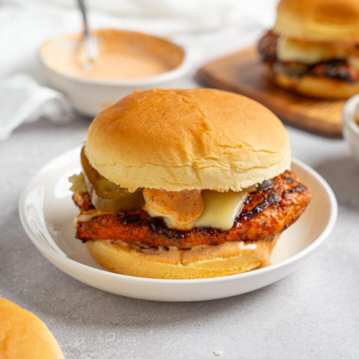 A homemade Popeye's blackened chicken sandwich on a white plate. The plate is on a white counter with a bowl of Popeye's sauce and an additional sandwich on a wooden board.
