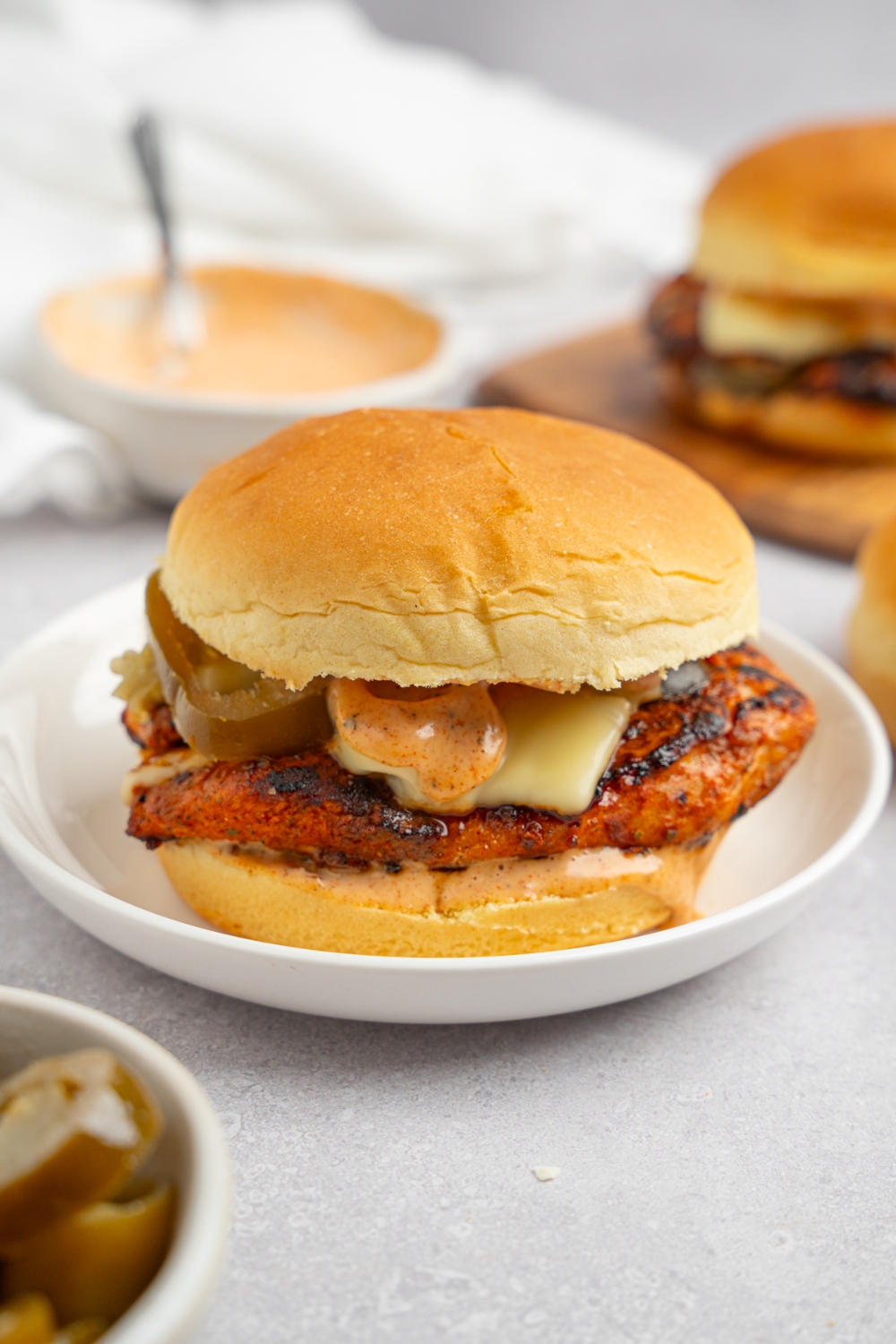 A homemade Popeye's blackened chicken sandwich on a white plate. The plate is on a white counter with a bowl of Popeye's sauce and an additional sandwich on a wooden board.
