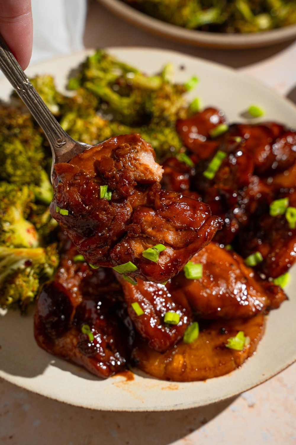 A close up of a fork with a bite of grilled Hawaiian BBQ chicken. There is a plate of chicken with roasted broccoli garnished with sliced green onion in the background.