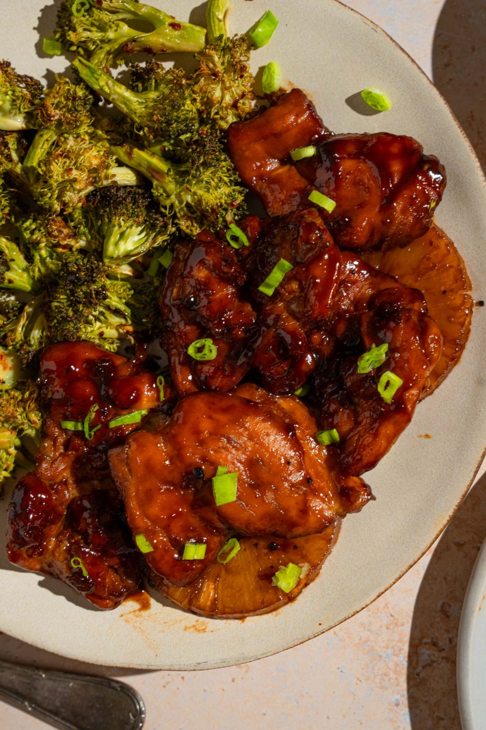 Grilled Hawaiian BBQ chicken on a white plate served with roasted broccoli and garnished with sliced green onion. The plate is on a tan counter.