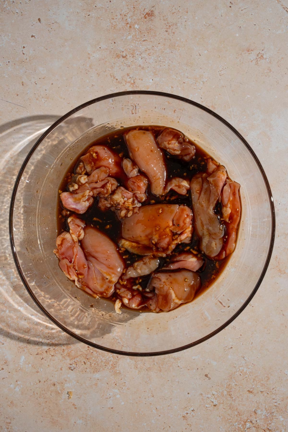 A glass bowl with chicken thighs marinating in a Hawaiian BBQ sauce. The bowl is on a tan counter.