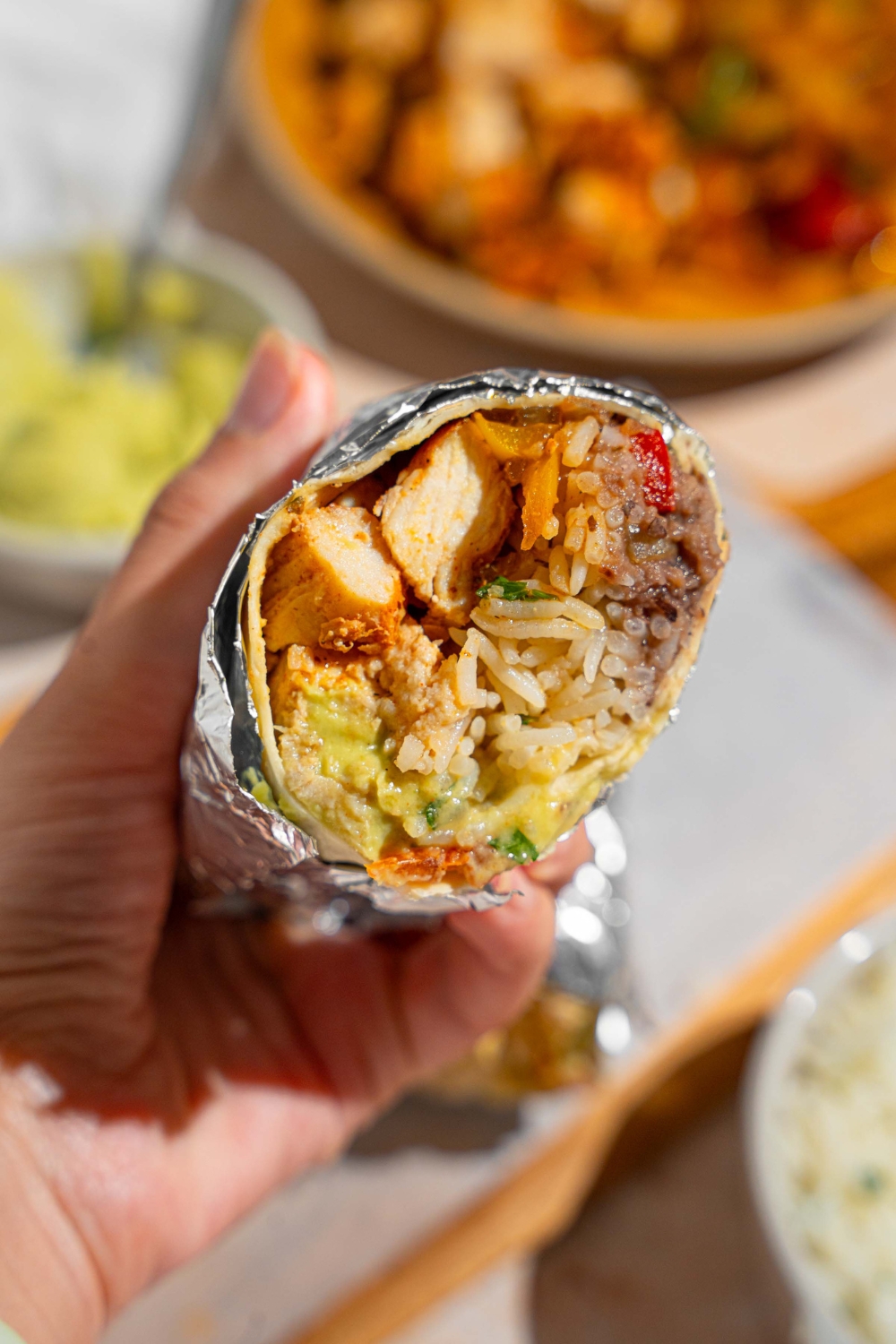A hand holding a half of a homemade Chipotle chicken burrito wrapped in foil. There is a wooden board with the other half of the burrito and bowls of burrito toppings blurred in the background.