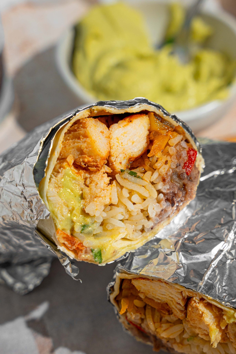A homemade Chipotle chicken burrito wrapped in foil and sliced in half. The burrito is on a wooden board lined with parchment paper. The board is next to a bowl of guacamole.