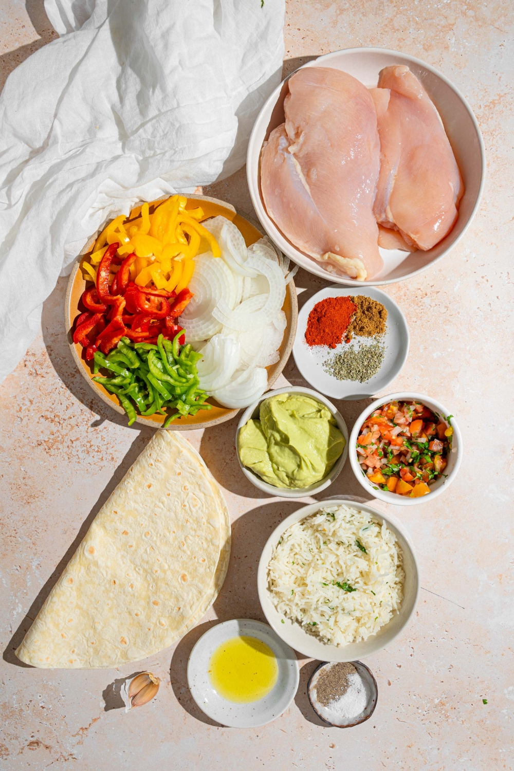 An overhead shot of several bowls in various sizes containing ingredients to make homemade Chipotle chicken burritos including tortilla wraps, chicken, cilantro lime rice, guacamole, salsa, garlic, sliced peppers and onions, and seasonings.