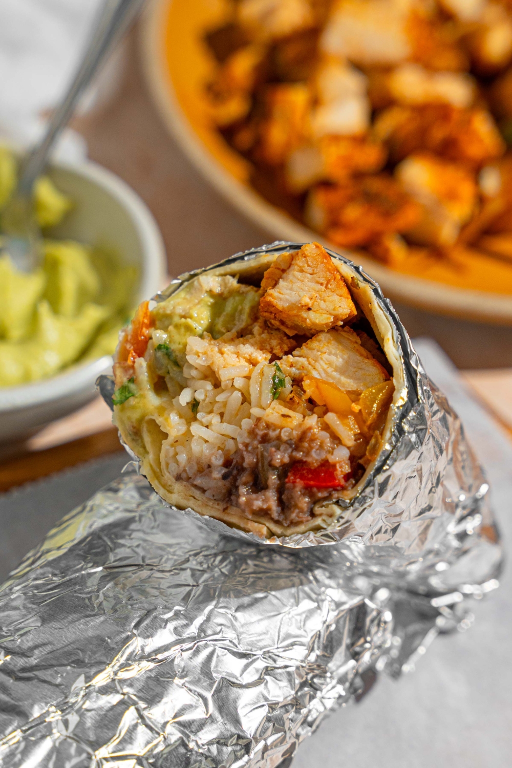 A homemade Chipotle chicken burrito sliced in half and wrapped in the foil. The halves are stacked on top of one another on a wooden board lined with parchment paper. The board is next to a plate of sautéed vegetables and bowl of guacamole.