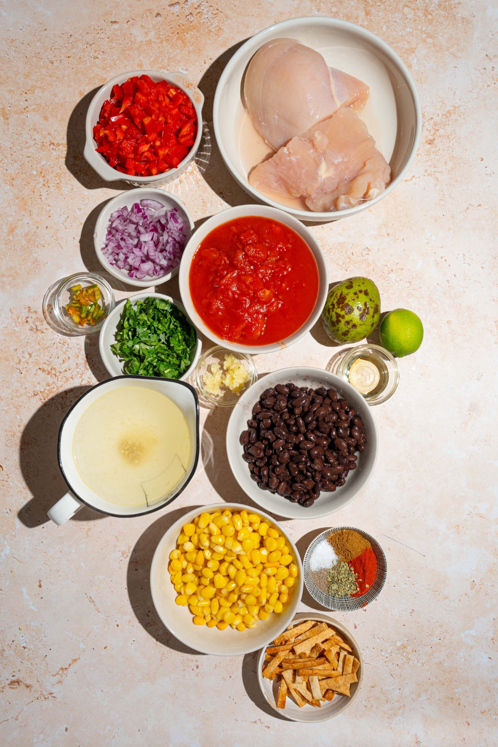 An overhead shot of several bowls in various sizes containing ingredients to make Panera chicken tortilla soup including chicken breasts, onion, corn, black beans, chicken broth, red pepper, tomatoes, jalapeno, lime, garlic, and seasonings.