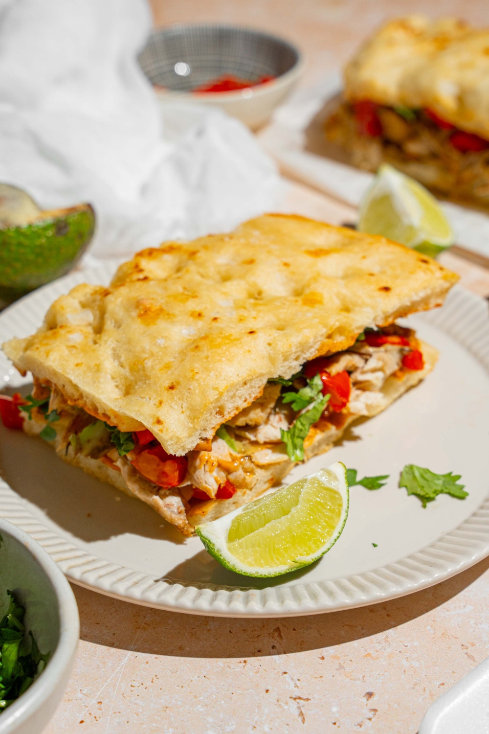 A white plate with a Panera chipotle chicken avocado melt garnished with fresh parsley and served with a lime wedge. The plate is on a tan counter with a small bowl of garnishes, white cloth napkin, and halved avocado.
