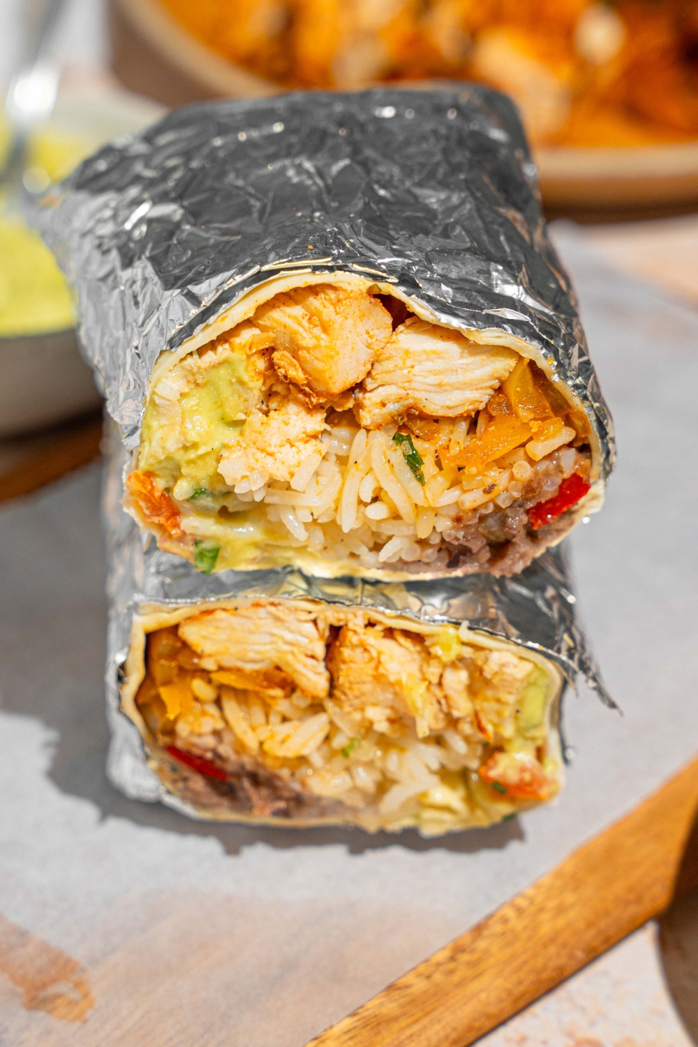 A homemade Chipotle chicken burrito sliced in half and wrapped in the foil. The halves are stacked on top of one another on a wooden board lined with parchment paper. The board is next to a plate of sautéed vegetables and bowl of guacamole.