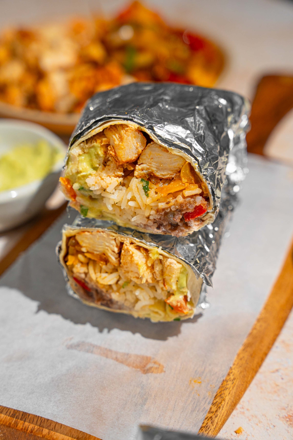 A homemade Chipotle chicken burrito sliced in half and wrapped in the foil. The halves are stacked on top of one another on a wooden board lined with parchment paper. The board is next to a plate of sautéed vegetables and bowl of guacamole.