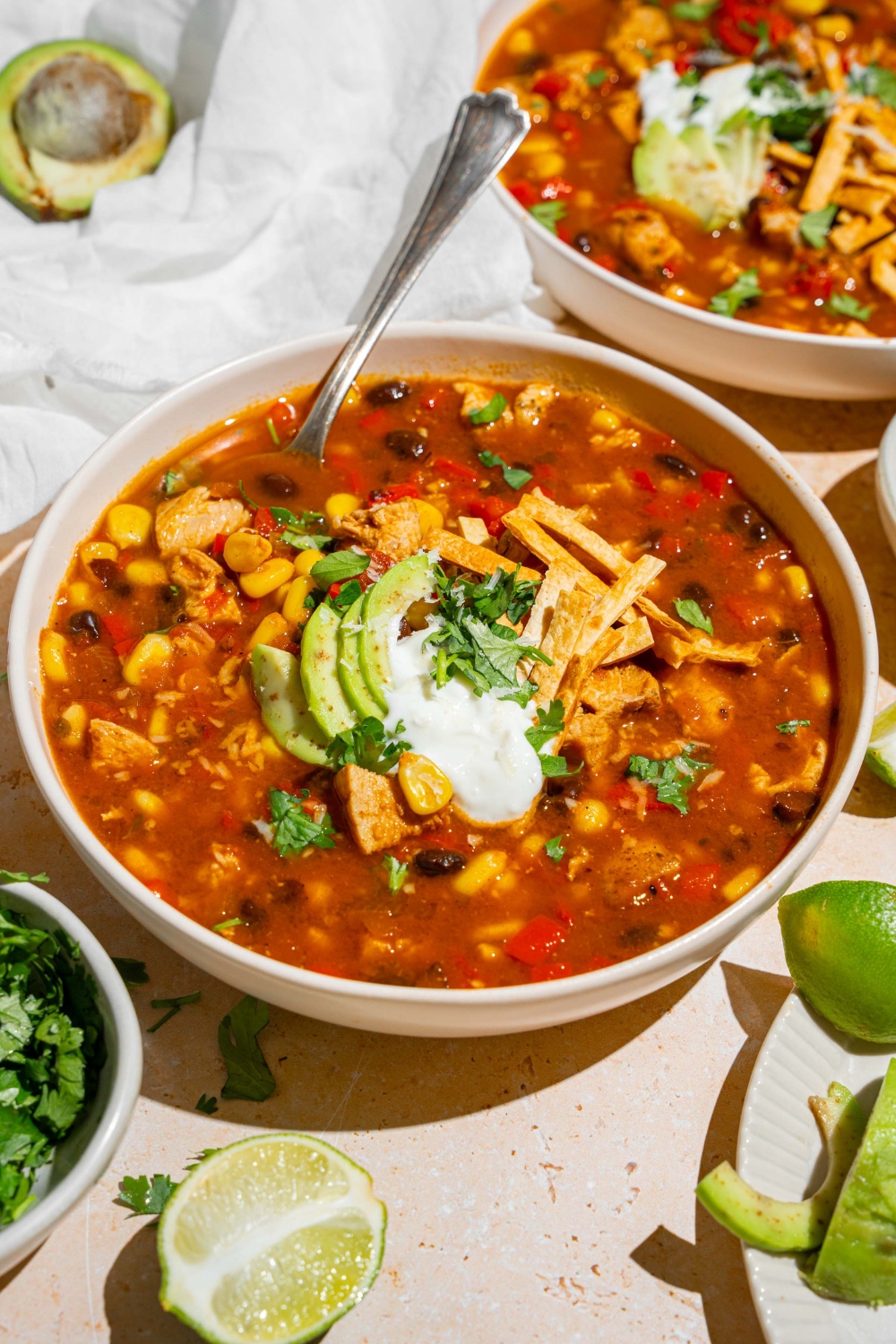 A white bowl with copycat Panera chicken tortilla soup topped with sour cream, sliced avocado, fresh cilantro, and tortilla strips. There is a spoon in the bowl. The bowl is on a tan counter with an additional bowl of soup and small plates of garnishes.