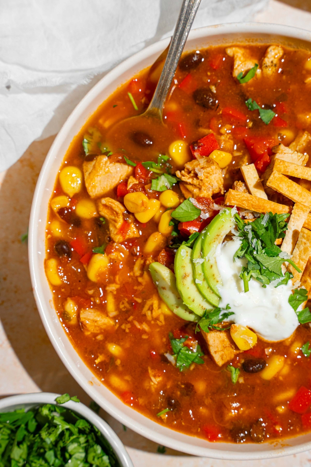 A white bowl with copycat Panera chicken tortilla soup topped with sour cream, sliced avocado, fresh cilantro, and tortilla strips. There is a spoon in the bowl. The bowl is on a tan counter with a small bowl of garnishes and white cloth napkin.