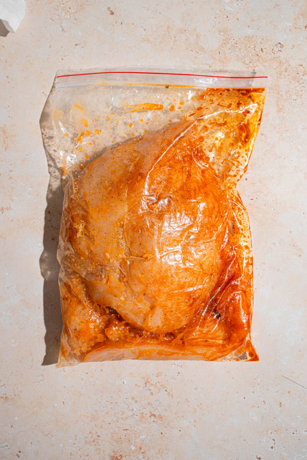 Chicken breasts marinating in a ziploc bag with seasonings. The bag is on a tan counter.