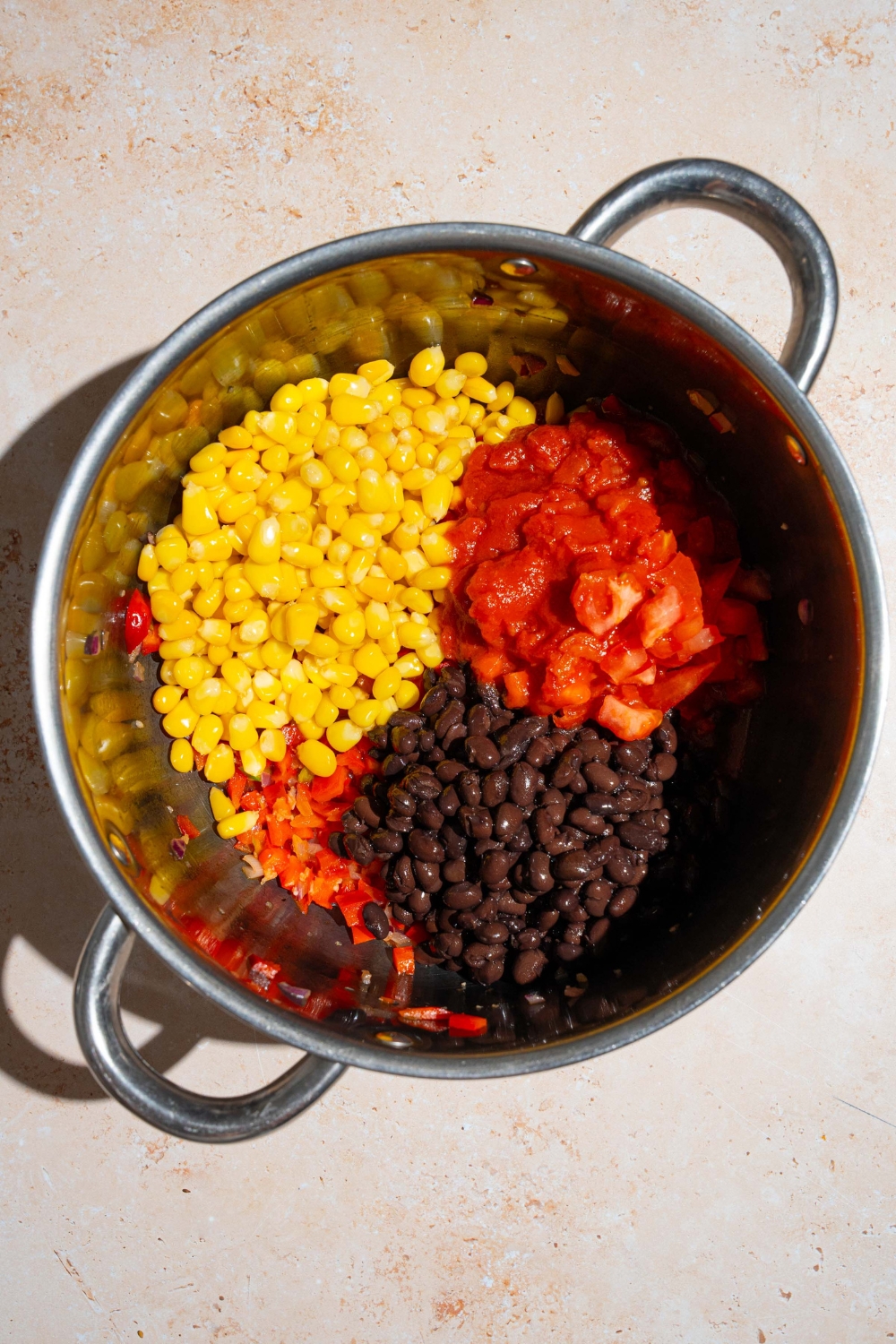 A stock pot with sautéed vegetables topped with corn, black beans, and diced tomatoes. The pot is on a tan counter.
