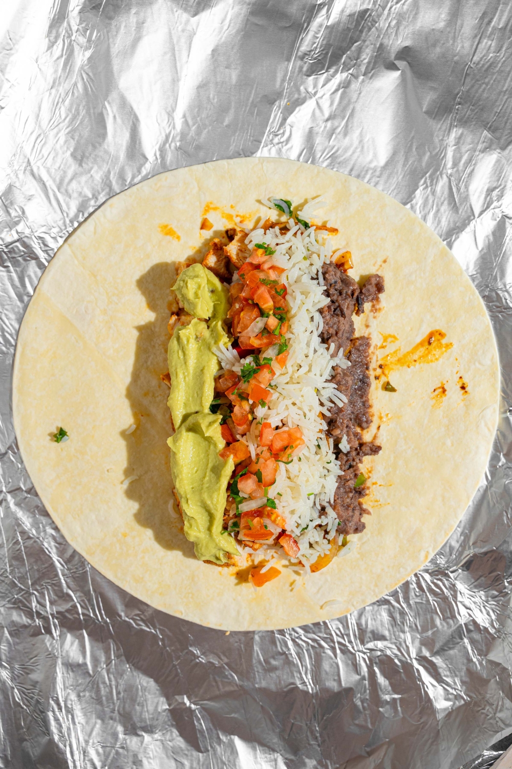 An open homemade Chipotle chicken burrito on a piece of foil. The tortilla is topped with chicken, sautéed vegetables, guacamole, salsa, black beans, and cheese.