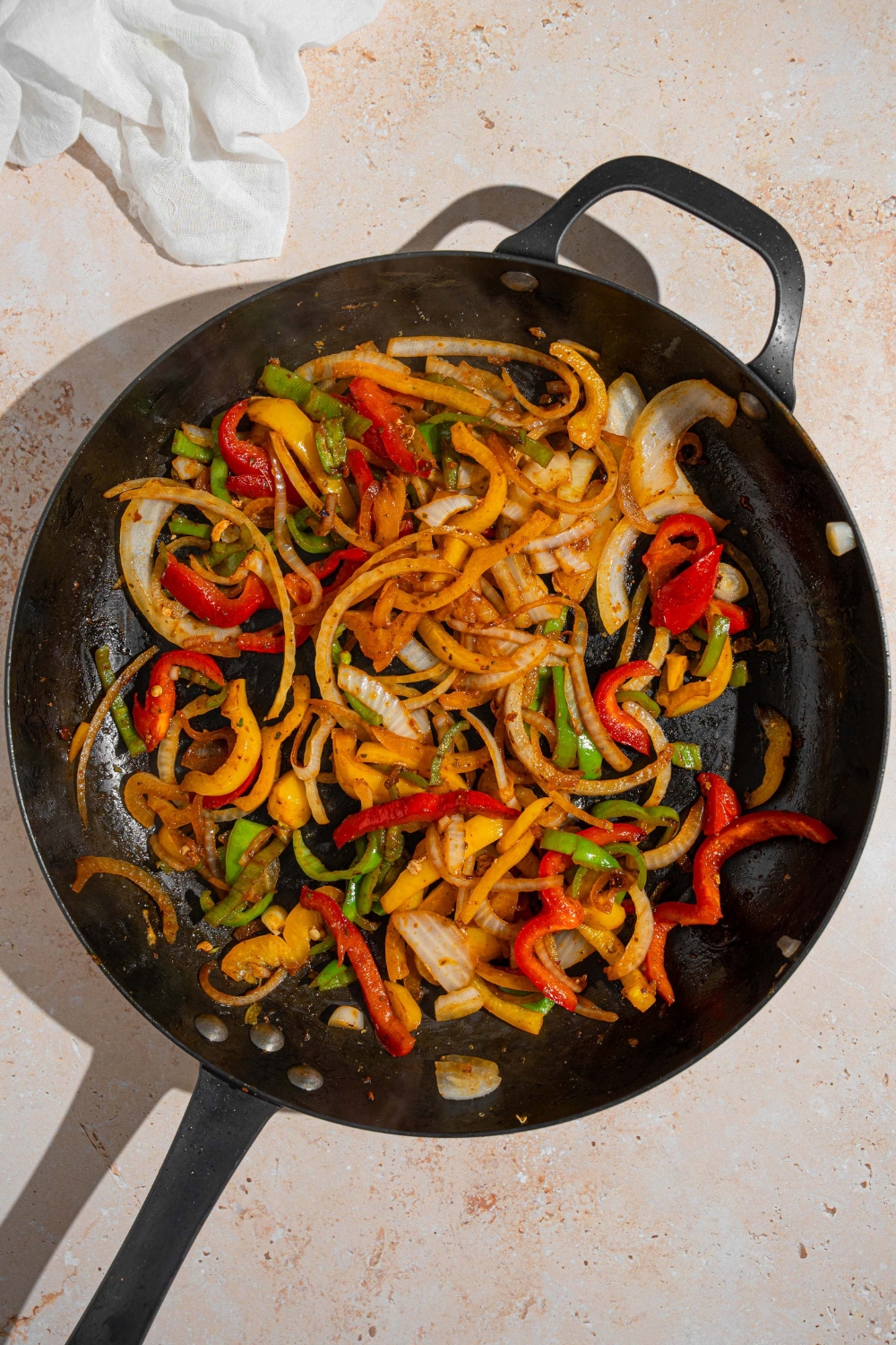 A cast iron skillet with sliced onion, red pepper, green pepper, and orange pepper sautéing in oil. The skillet is on a tan counter with a white cloth napkin.
