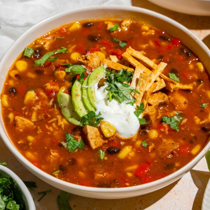 A white bowl with copycat Panera chicken tortilla soup topped with sour cream, sliced avocado, fresh cilantro, and tortilla strips. The bowl is on a tan counter with an additional bowl of soup and white cloth napkin.