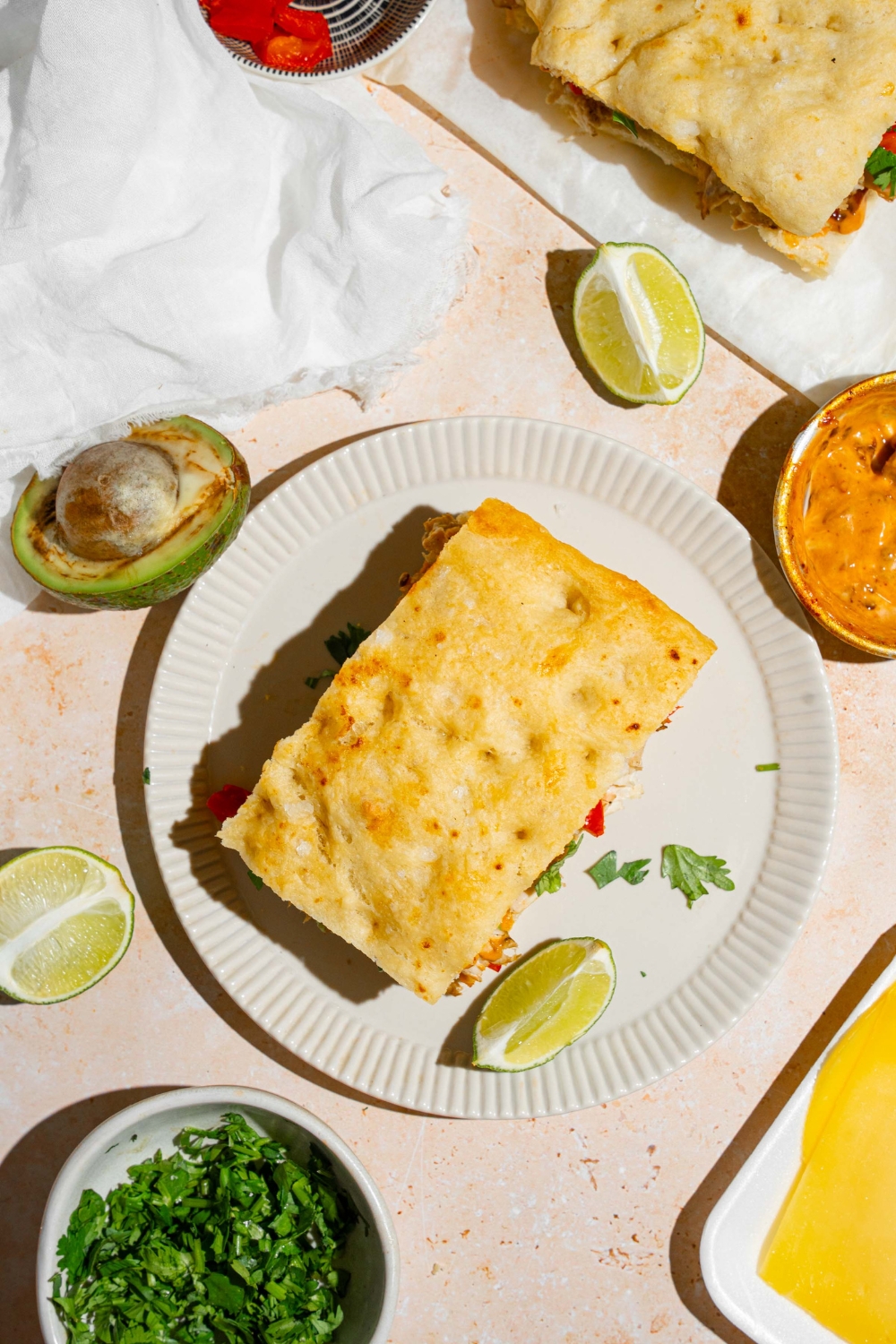 A white plate with a Panera chipotle chicken avocado melt garnished with fresh parsley and served with a lime wedge. The plate is on a tan counter with a small bowl of garnishes, white cloth napkin, and halved avocado.