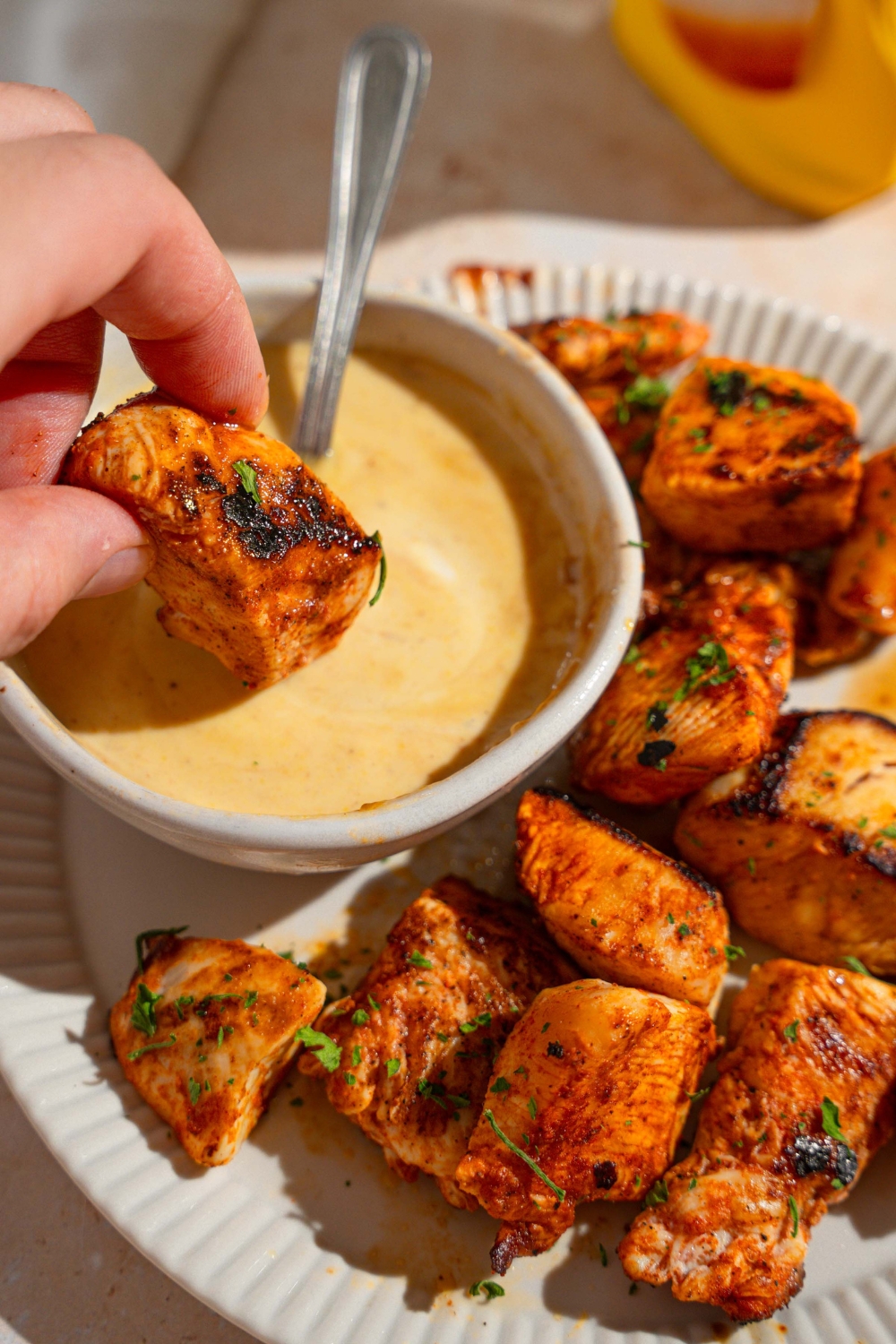 A hand dipping a copycat Chick Fil A nugget in a bowl of dipping sauce with a spoon in the bowl. The bowl is on a plate with additional nuggets.