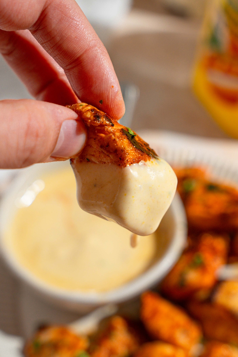 A close up of a hand holding a copycat Chick Fil A nugget dipped in dipping sauce. There is a plate of nuggets with a bowl of sauce blurred in the background.