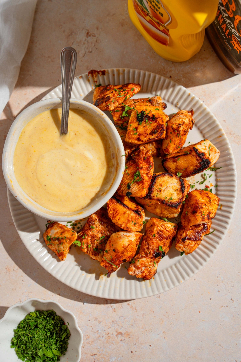 A white plate with homemade Chick Fil A nuggets garnished with fresh parsley. There is a bowl of dipping sauce on the plate with a spoon in the sauce. The plate is on a tan counter with a white cloth napkin and condiments.