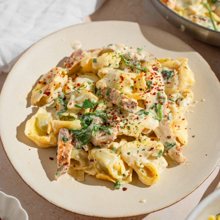 A white plate with copycat Olive Garden chicken tortellini alfredo garnished with red pepper flakes. The plate is on a tan counter with a skillet of chicken alfredo and a white cloth napkin.