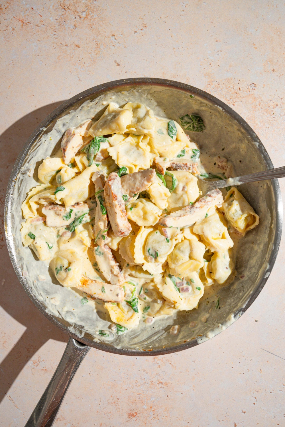 A skillet with a spoon stirring copycat Olive Garden chicken tortellini alfredo. The skillet is on a tan counter.