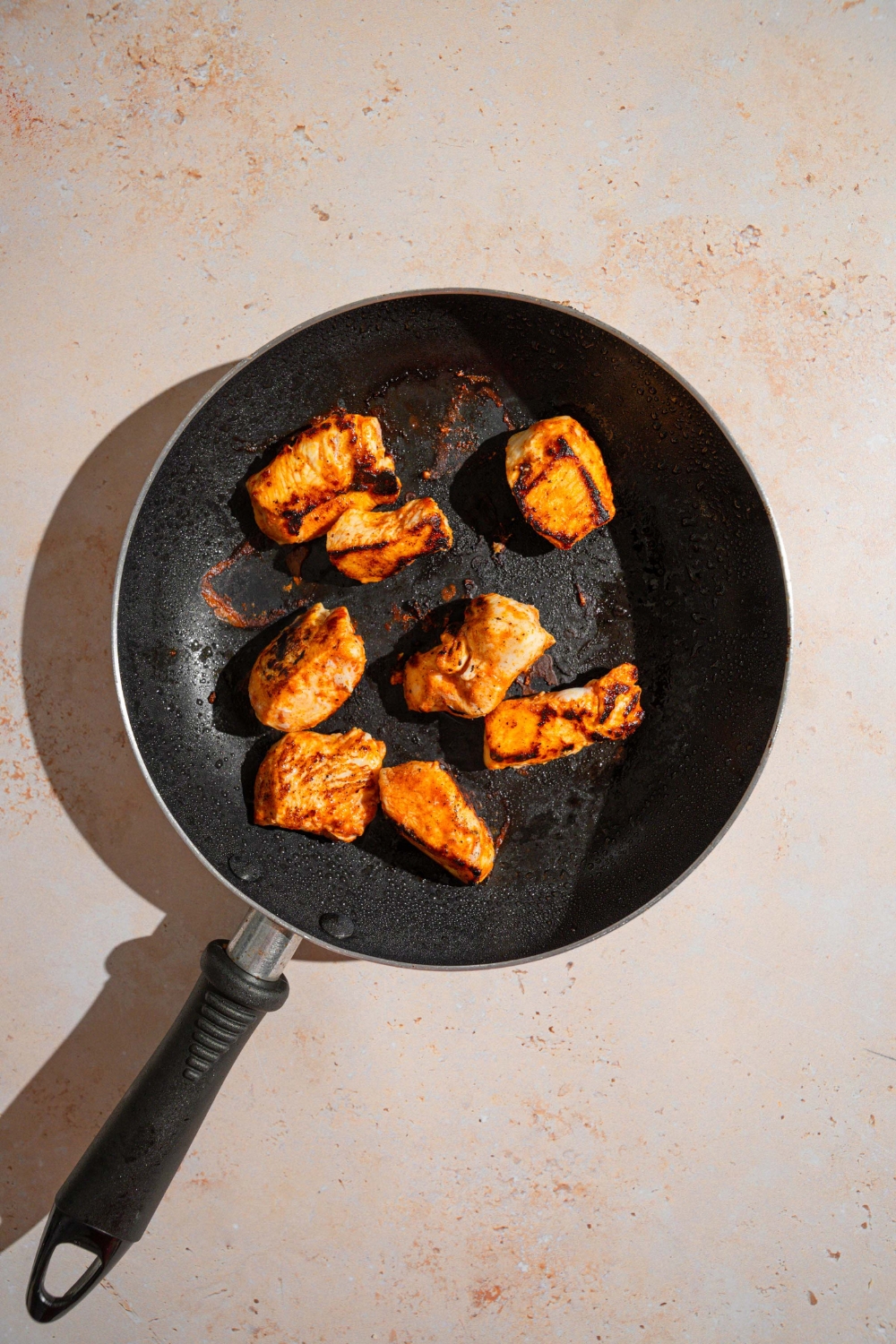 A skillet with several seasoned chicken breasts cooking in oil. The skillet is on a tan counter.